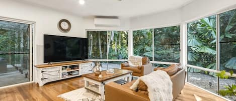 A modern living room with a flat-screen TV, wooden flooring, a ceiling fan, and large windows offering a view of greenery.