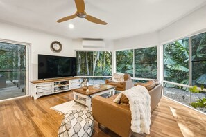 Ein modernes Wohnzimmer mit einem Flachbildfernseher, Holzboden, einem Deckenventilator und großen Fenstern mit Blick auf Grünflächen.