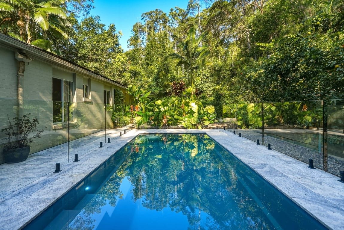 A rectangular swimming pool with clear water, surrounded by a white tiled deck, and bordered by a glass fence.