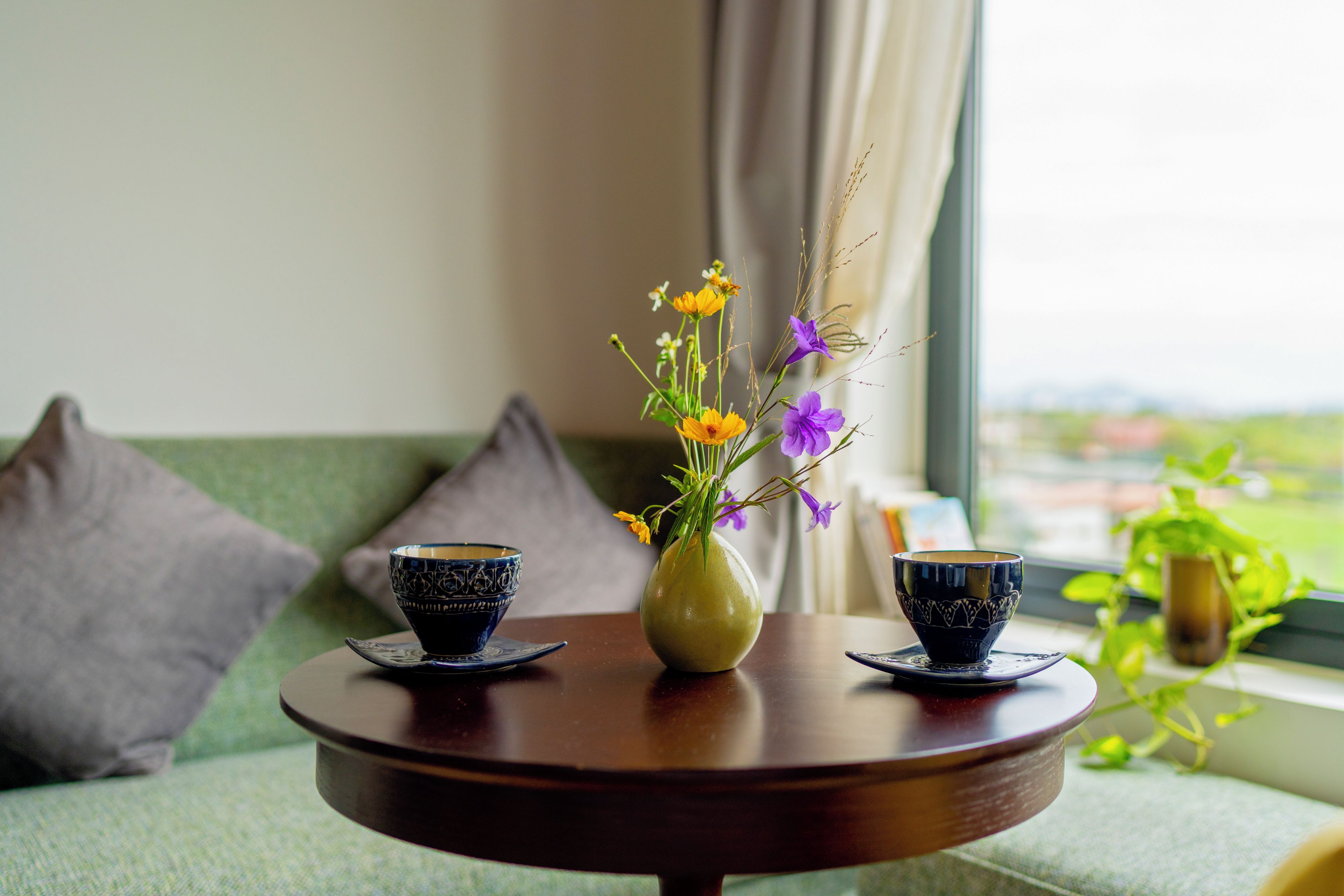 Una mesa redonda de madera con dos tazas y platillos de diseño oscuro, un jarrón amarillo con flores, y una vista de un paisaje verde a través de una ventana.