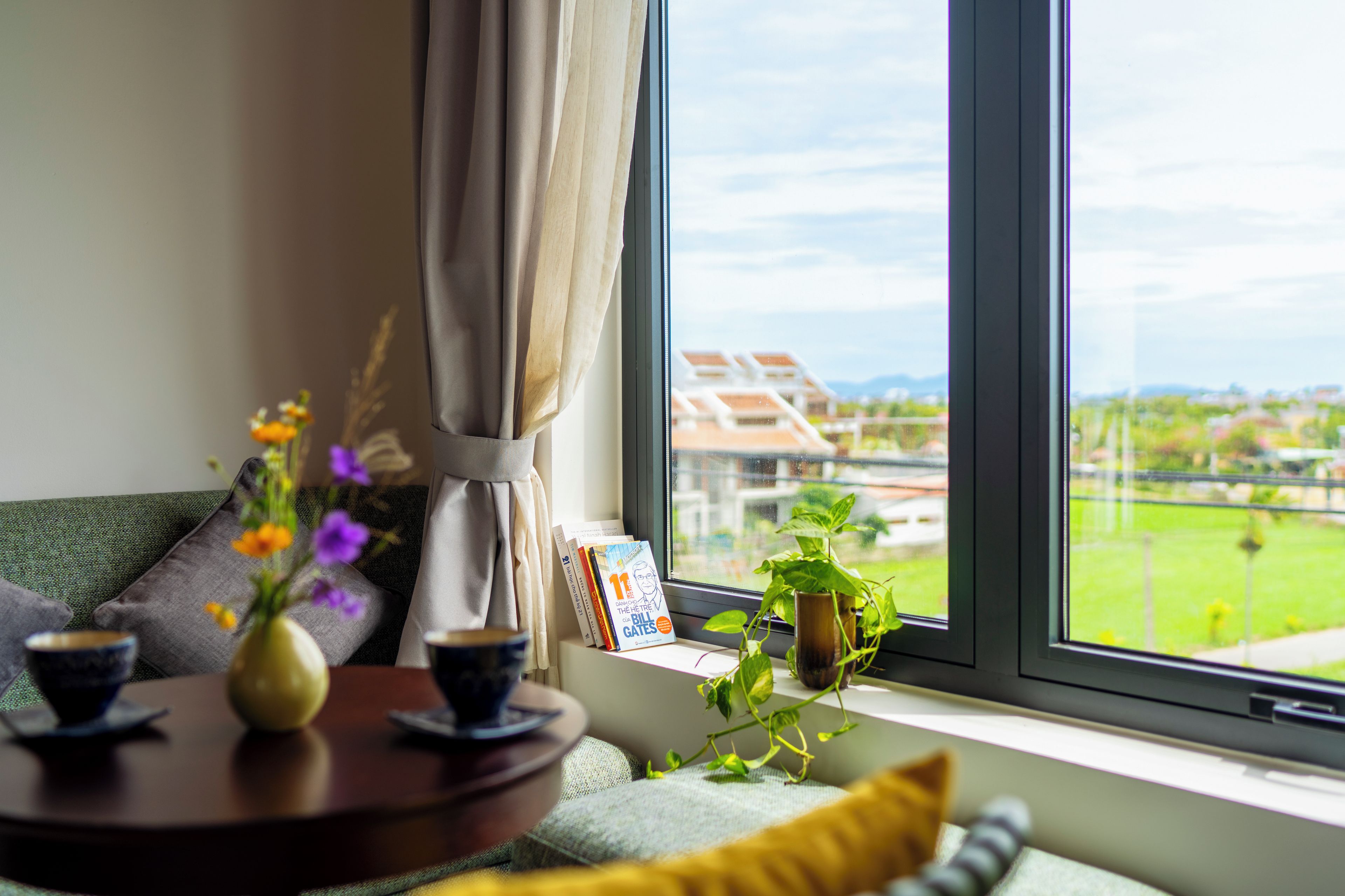 Una habitación con vistas a un campo verde y edificios, una ventana con cortina, una mesa con un jarrón de flores y una estantería con libros.