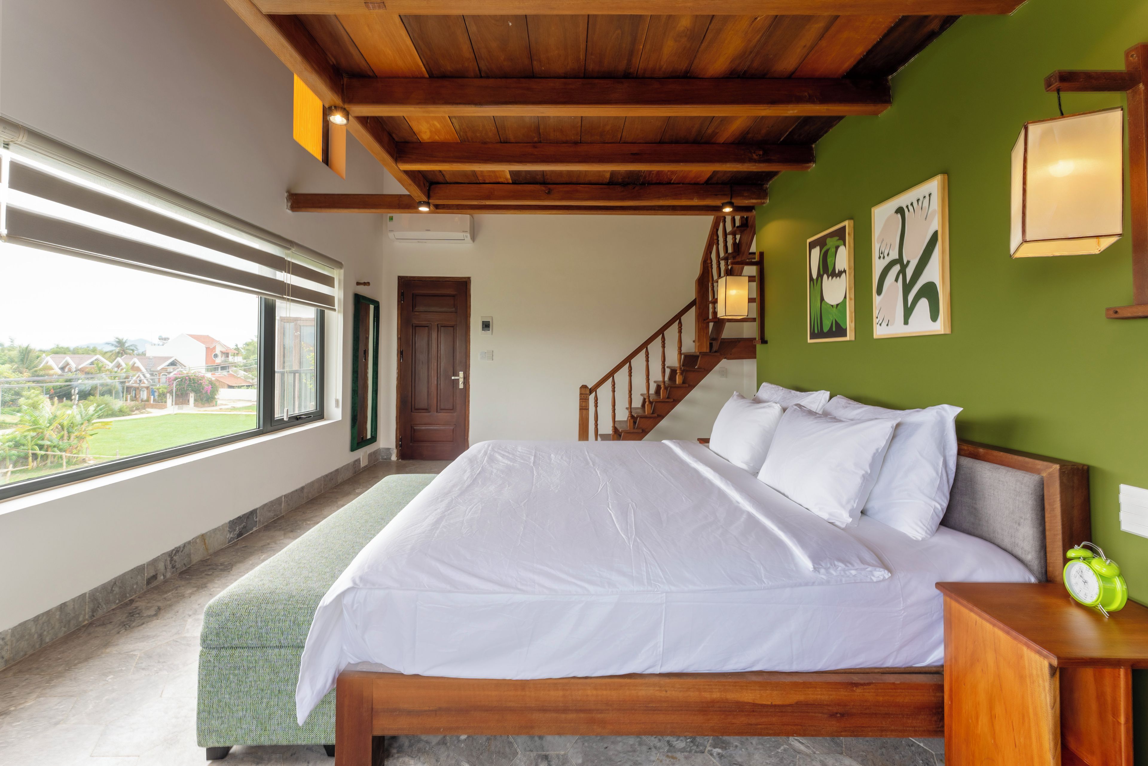 Un dormitorio con una cama grande, cabecera de madera, pared verde, una escalera y una ventana con vistas al exterior.