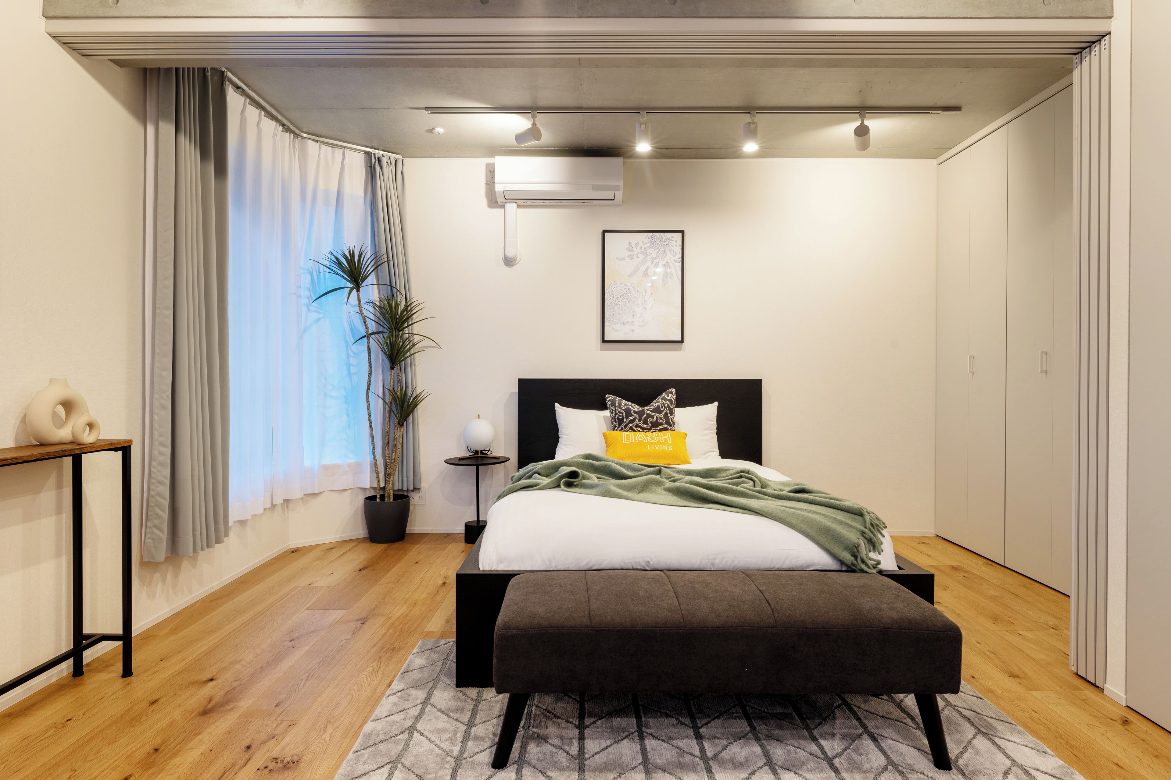 A modern bedroom with a bed, bedside table, potted plants, and a window with curtains.
