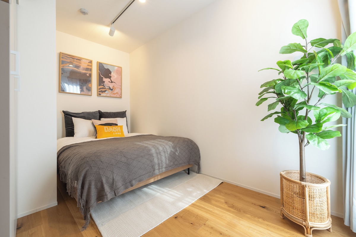 Quarto com cama, manta cinzenta, tapete branco, planta numa cesta de vime e duas pinturas abstratas na parede.