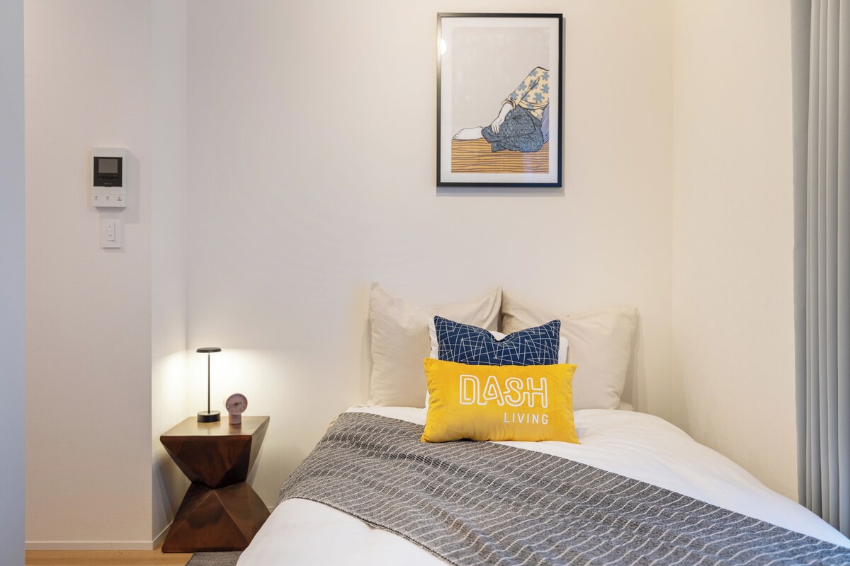 Quarto de hotel com cama, mesa de cabeceira com abajur, termostato fixado na parede e uma obra de arte emoldurada acima da cama.