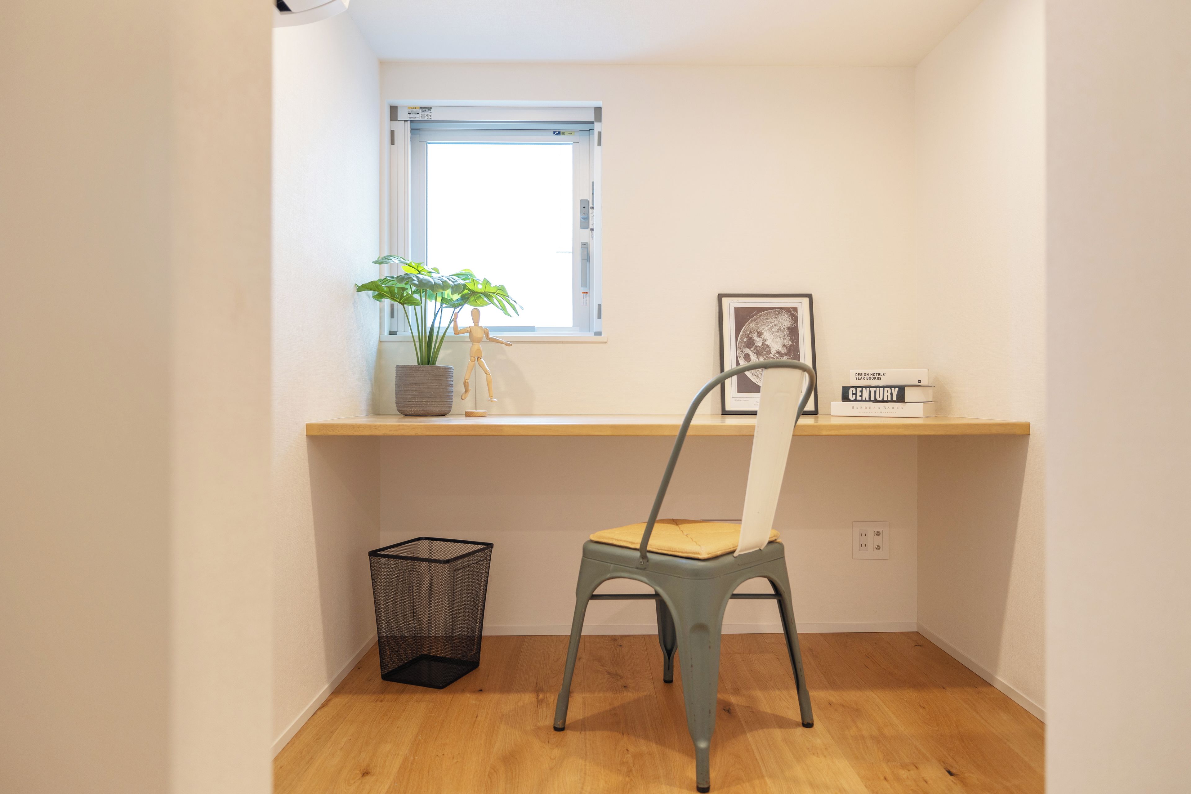 Een kleine kamer met een bureau, een stoel, een raam, een plant en een lijst met een afbeelding.