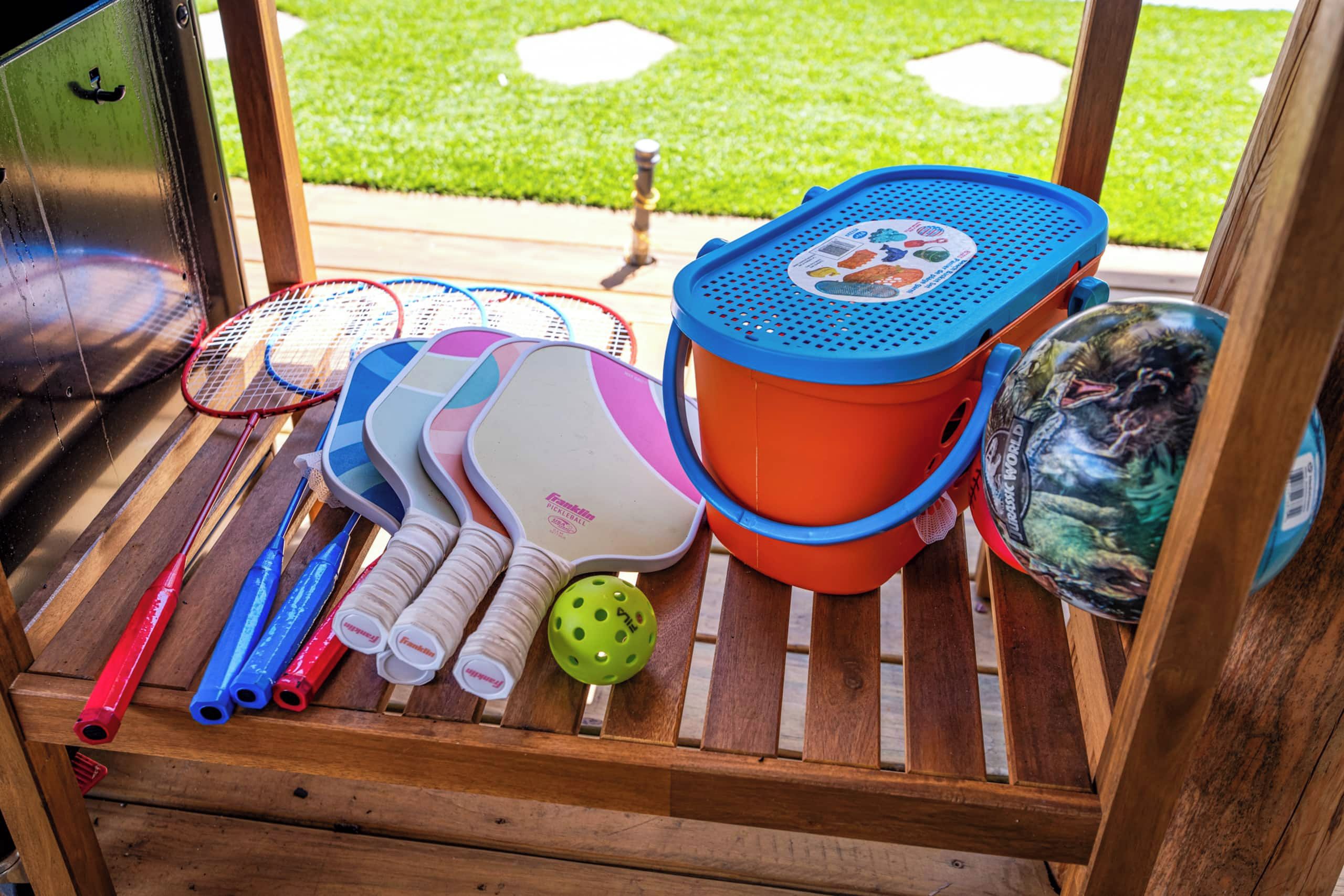 A wooden bench with various sports equipment including badminton rackets, a pickleball paddle, and a croquet ball.