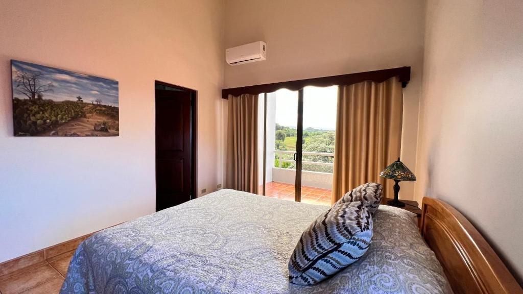 Een slaapkamer met een bed, een schilderij, een raam met gordijnen en uitzicht op een landschap.