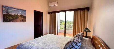 A bedroom with a bed, a painting, a window with curtains, and a view of a landscape.
