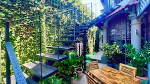 Un patio exterior con una mesa y sillas de madera, rodeado de abundante vegetación y una pérgola de metal.