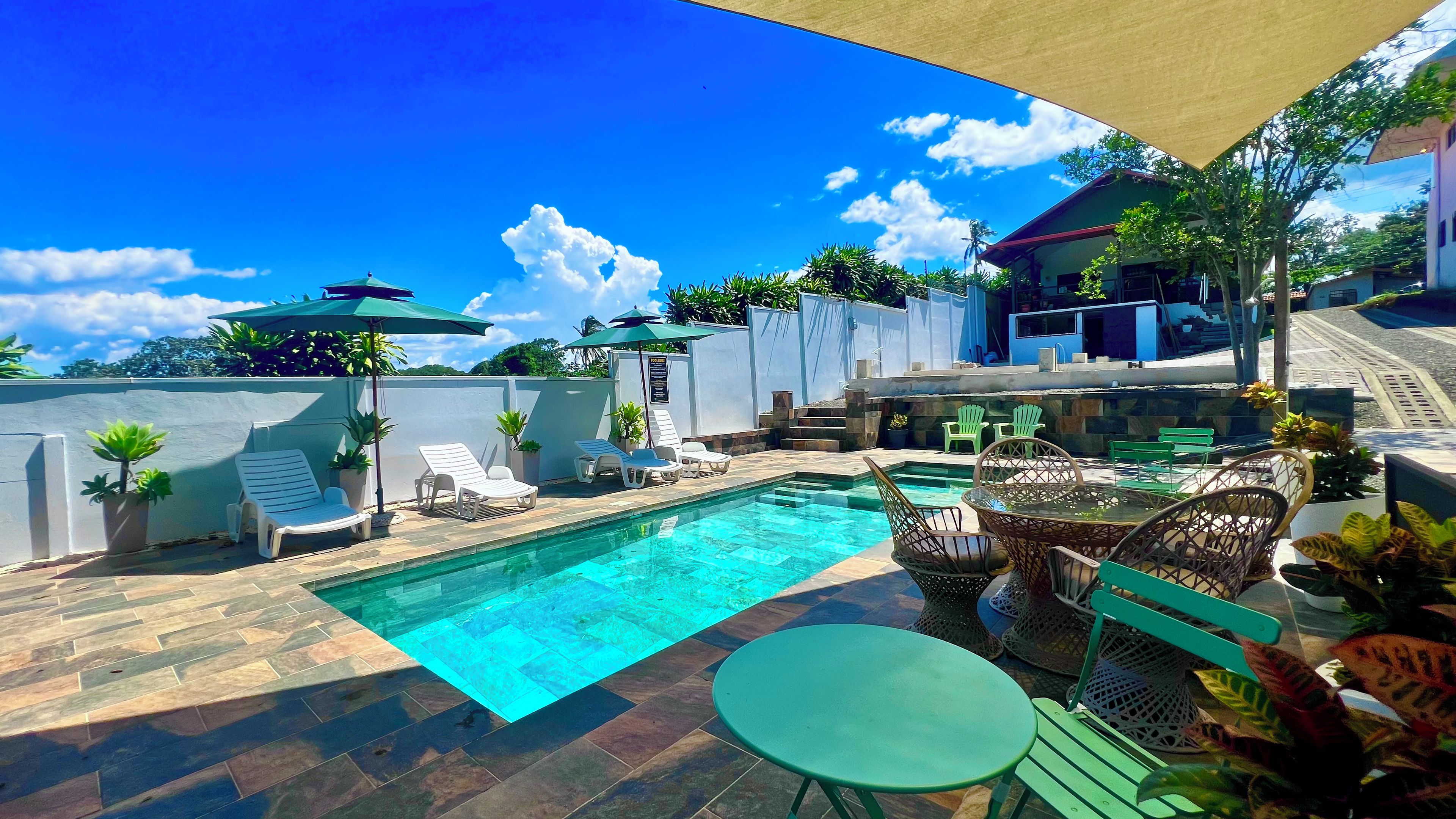 Piscina com espreguiçadeiras e uma mesa, rodeada por plantas em vasos e uma área sombreada.