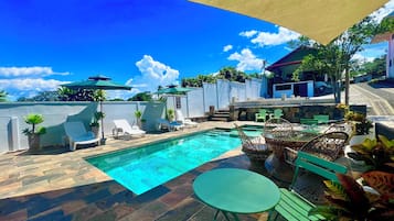 Piscina com espreguiçadeiras e uma mesa, rodeada por plantas em vasos e uma área sombreada.