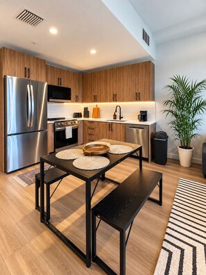 A modern kitchen with wooden cabinets, stainless steel appliances, and a dining area with a black table and bench. - Close to Beach | Stylish 1BR + Pullout Sofa (Hollywood)