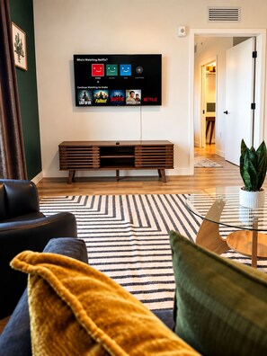 Ein modernes Wohnzimmer mit einem Flachbildfernseher, der eine Netflix-Oberfläche anzeigt, einer Holzbank, einem Sofa und einem gemusterten Teppich.