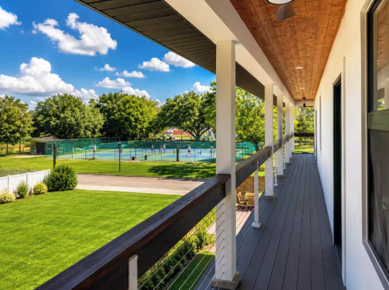 A covered outdoor area with a wooden deck, white railings, and a view of a tennis court.