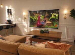 Ein Wohnzimmer mit einem großen Flachbildfernseher an der Wand, einem Holztisch und einem beigefarbenen Sofa.