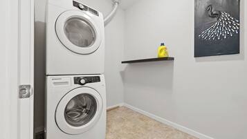 A laundry room with two white Samsung front-loading washers, a yellow detergent bottle on a shelf, and a black abstract painting on the wall.