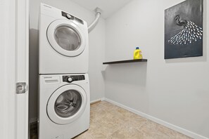 Une buanderie équipée de deux lave-linge Samsung à chargement frontal blancs, d’une bouteille de lessive jaune posée sur une étagère et d’un tableau abstrait noir accroché au mur.