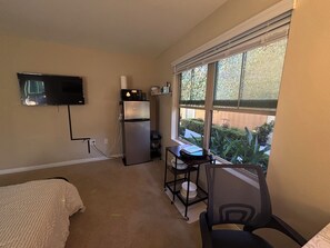Habitación compacta con cama, refrigerador, televisión montada en la pared y ventana con vista a la vegetación.