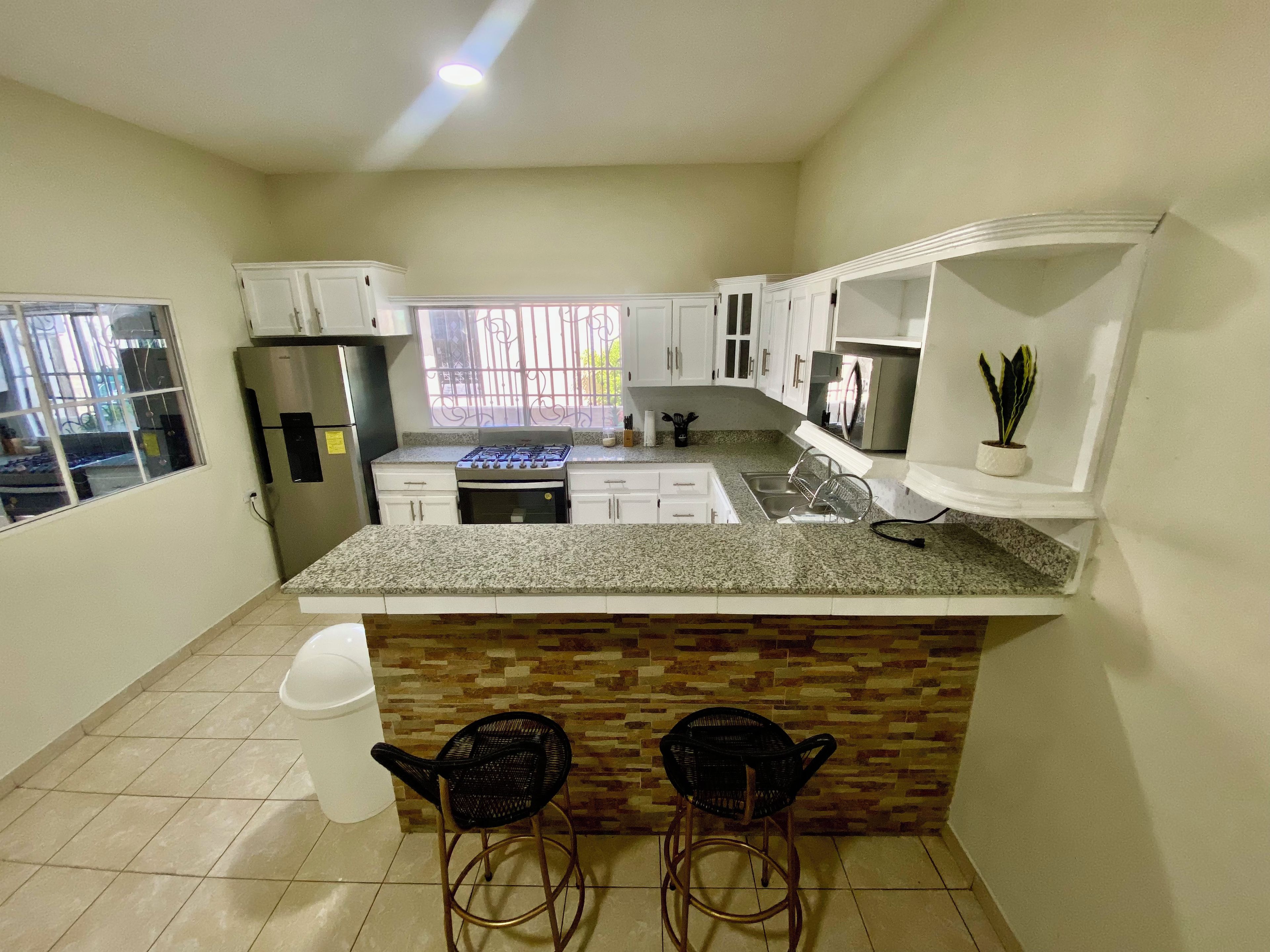 A kitchen with granite countertops, white cabinets, a stainless steel refrigerator, a gas stove, and a sink. There are two black chairs at the island.