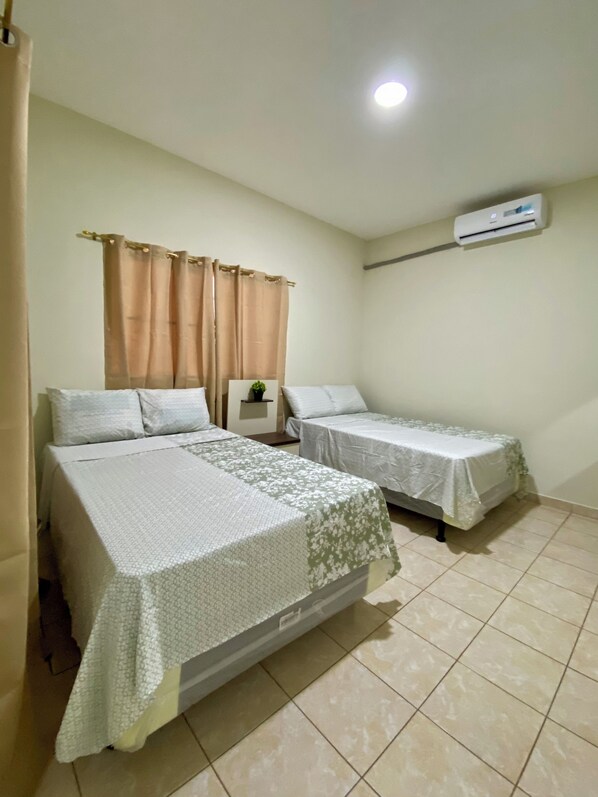 A hotel room with two beds, a small bedside table with a plant, beige curtains, an air conditioning unit, and a tiled floor.