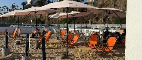 A beachside area with lounge chairs and umbrellas, a sign indicating no alcohol beyond a certain point, and a view of the ocean and hills in the background.