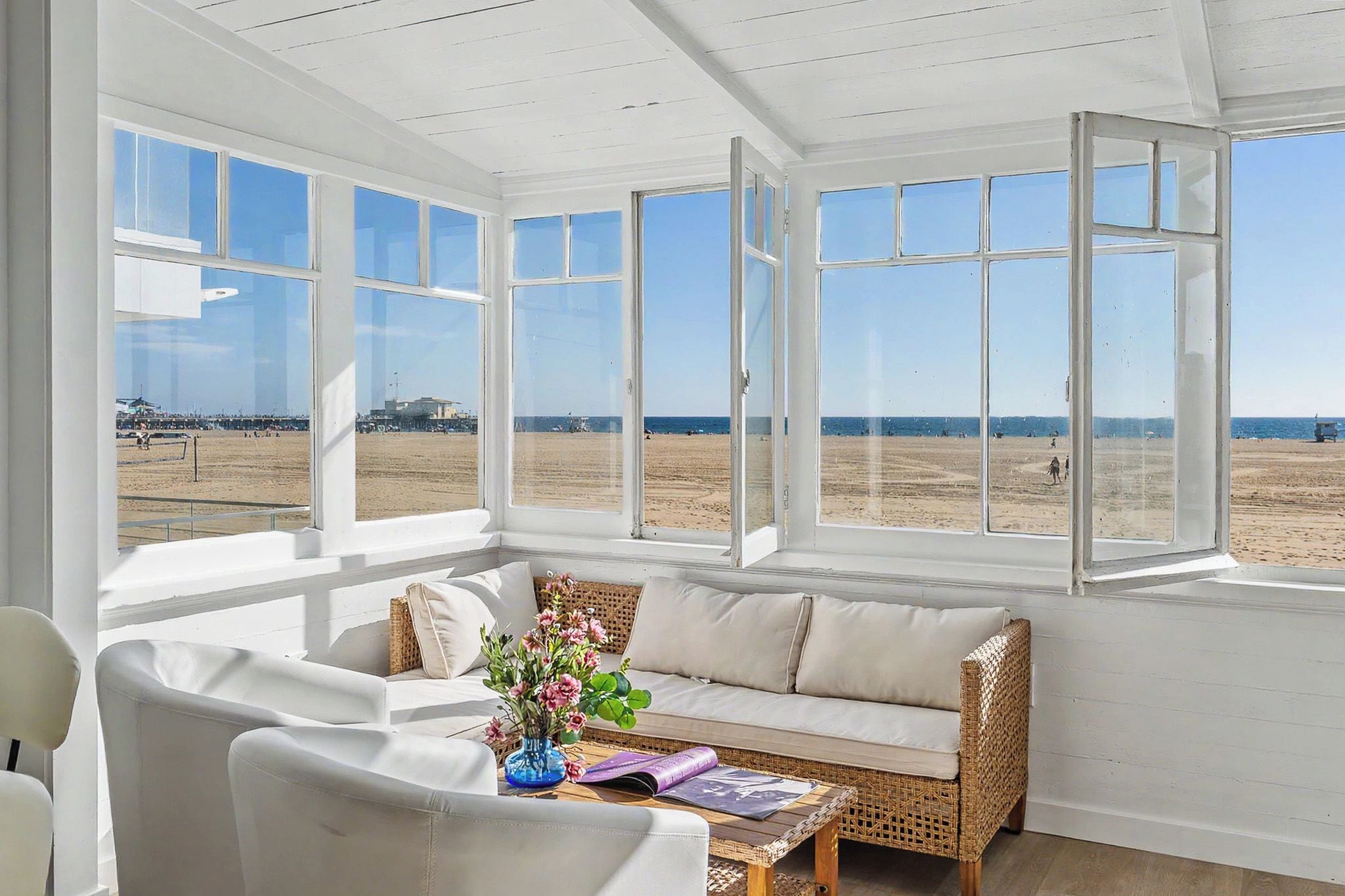 A sunlit room with a white sofa, wicker furniture, and a view of the beach.