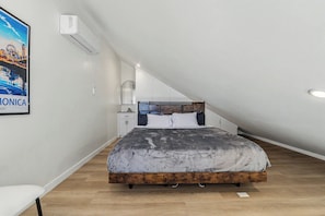 A bedroom with a sloped ceiling, a bed with a wooden frame, a nightstand, a wall-mounted air conditioner, and a poster on the wall.