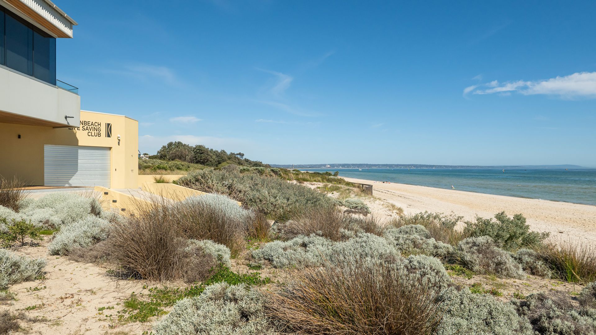 Ein modernes Gebäude mit einem Schild, auf dem 'NBEACH LIFE SAVING CLUB' steht, neben einem sandigen Strand mit spärlicher Vegetation.