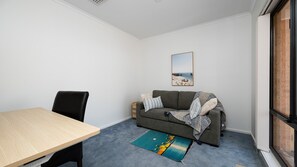A room with a wooden table, a grey sofa with a blanket, a framed picture on the wall, and a window with a brick wall view.