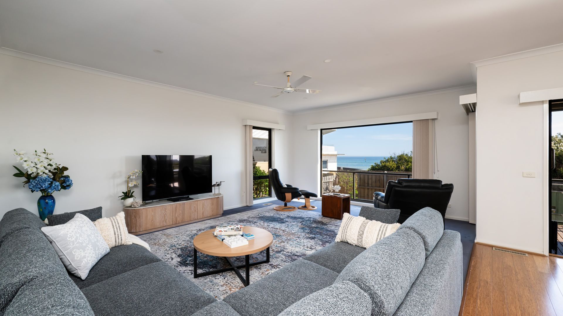 A modern living room with a flat-screen TV, grey sofas, a coffee table, and a view of the ocean.