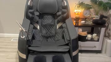 Een massagestoel met voetsteun en bedieningsknoppen, staand in een ruimte met een bijzettafeltje waarop een lamp, een klok en een speaker staan.