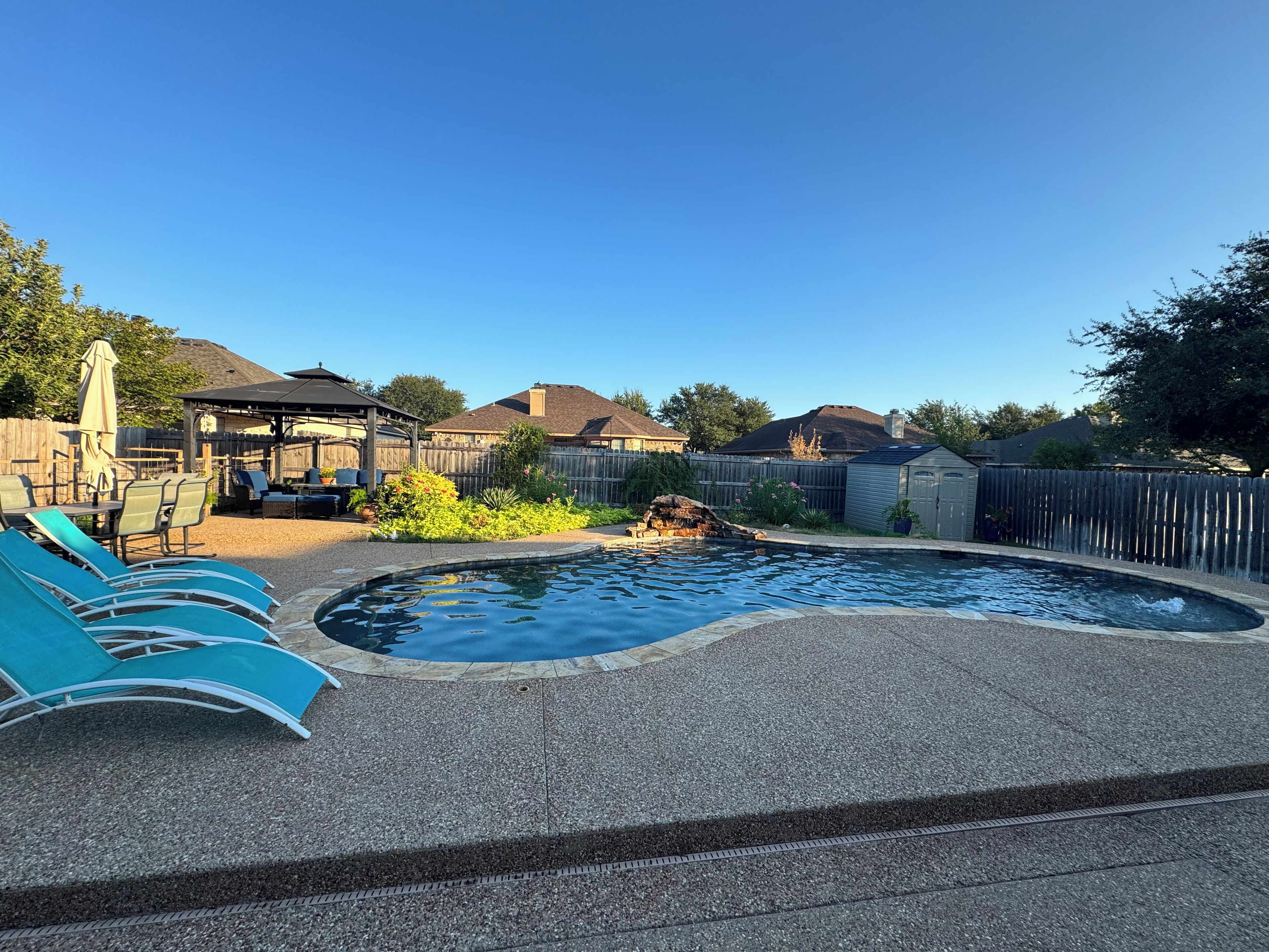 Un jardin avec une piscine, des chaises longues, une terrasse couverte et une clôture en bois.