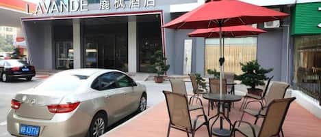 Entrada de hotel con un coche estacionado frente, mesas al aire libre con un paraguas rojo y un letrero.
