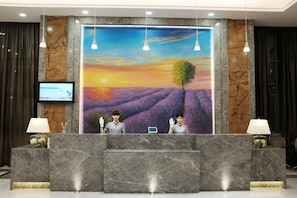 A reception desk with a large mural of a sunset over a lavender field, two receptionists, and a computer monitor. - Lavande Hotel (Dongguan)