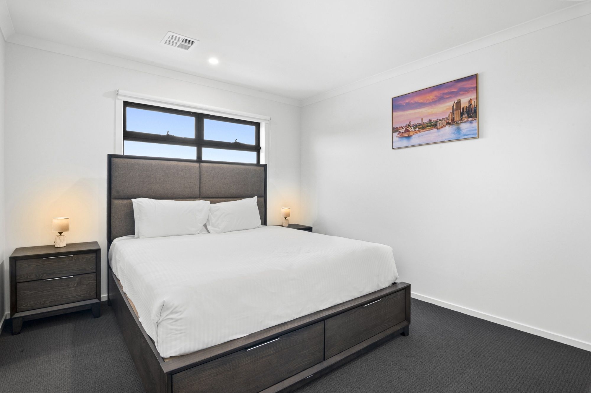 A bedroom with a bed, two bedside tables, a window, and a wall-mounted picture.