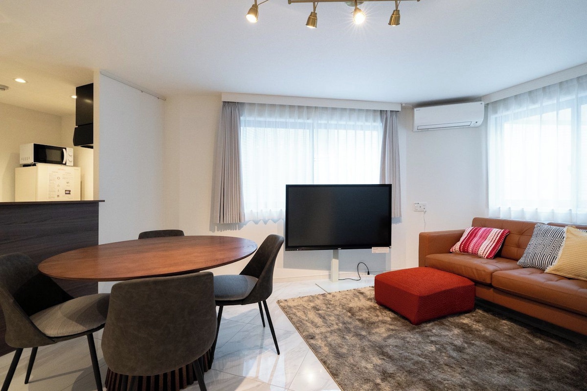 Ein modernes Wohnzimmer mit einem Flachbildfernseher, einem runden Esstisch mit Stühlen, einer Couch mit Kissen und einem großen Fenster mit Vorhängen.