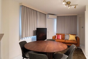 Ein modernes Wohnzimmer mit einem runden Holztisch, grau gepolsterten Sesseln, einem braunen Sofa mit gemusterten Kissen und einer Deckenleuchte.