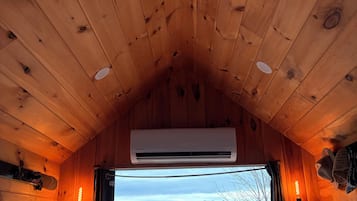 A cabin interior with wooden walls, a sloped ceiling, an air conditioning unit, a window with curtains, and a view of a snowy landscape.