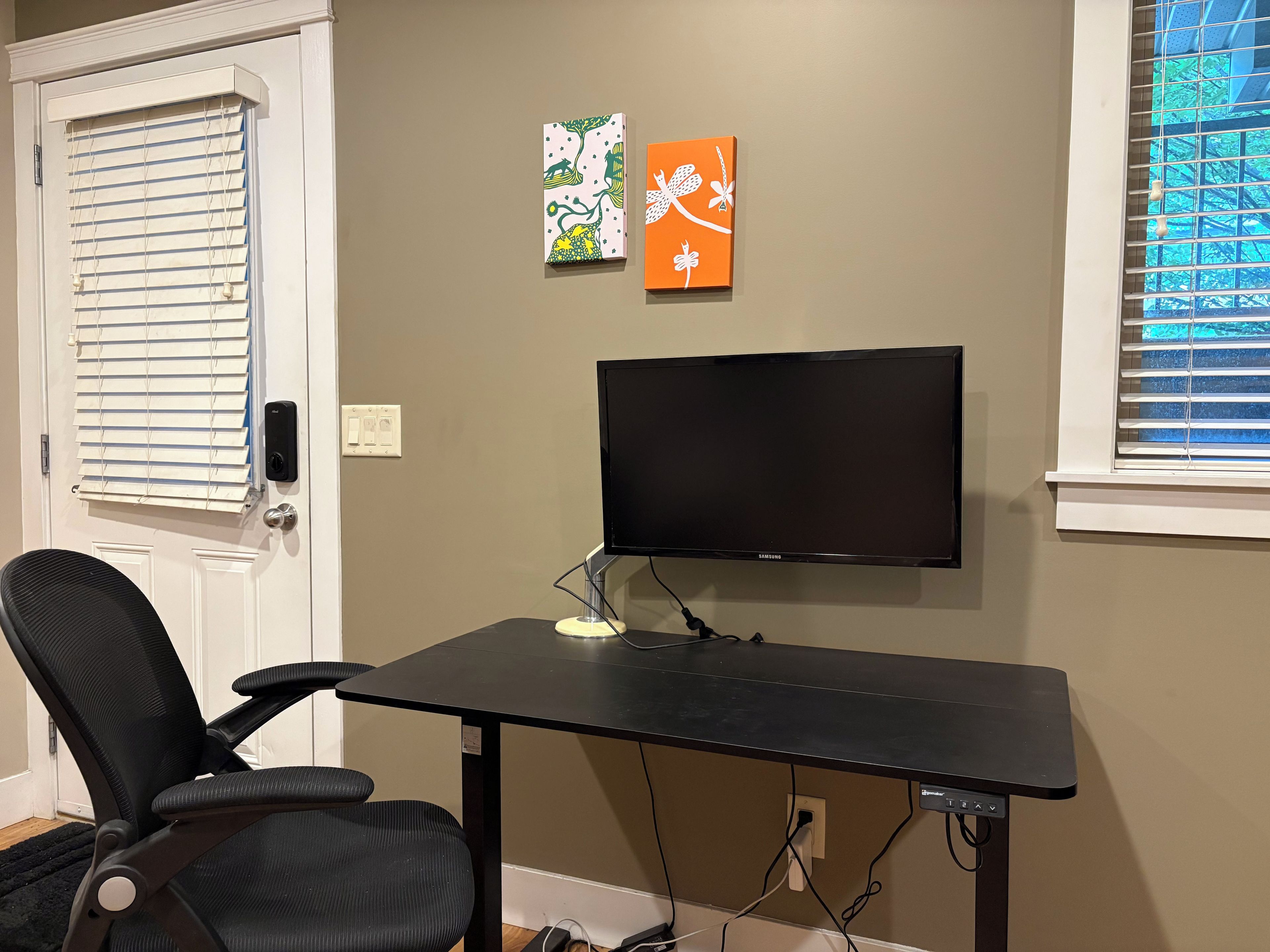 A room with a black desk, a black office chair, a mounted flat-screen TV, and two wall art pieces.
