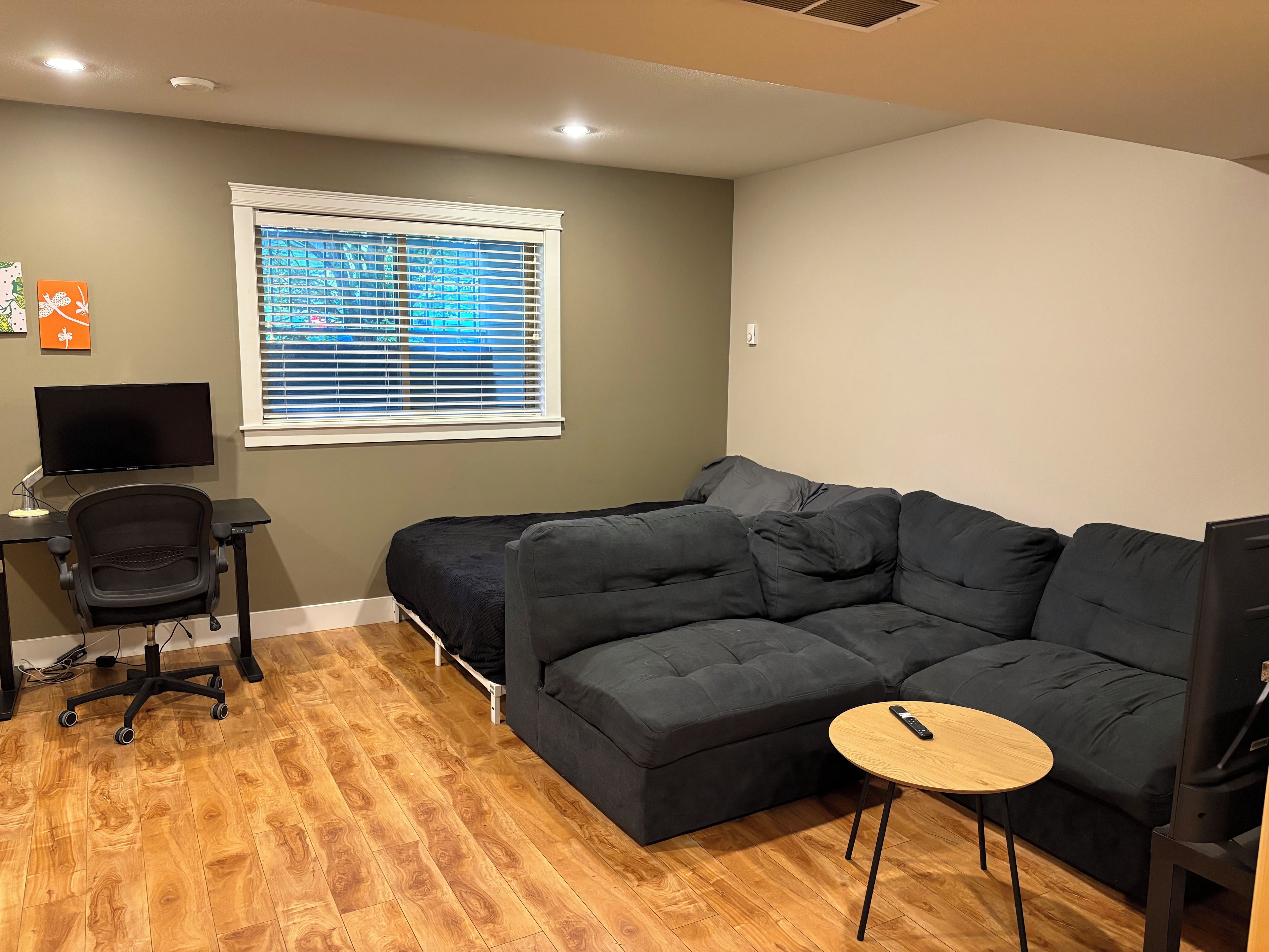 Una habitación con un sofá modular, un escritorio con una computadora y una ventana con persianas.