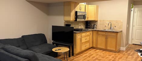 A compact living space with a sofa, a television, a kitchen area with wooden cabinets and a microwave, and a small round table.