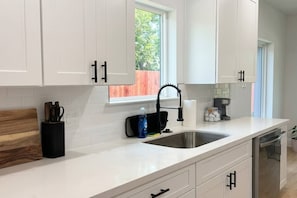 A modern kitchen with white cabinets, a stainless steel sink, and a window with a view of greenery. - Modern 2BR Townhome | Medical Center • NRG • Hobby (Houston)