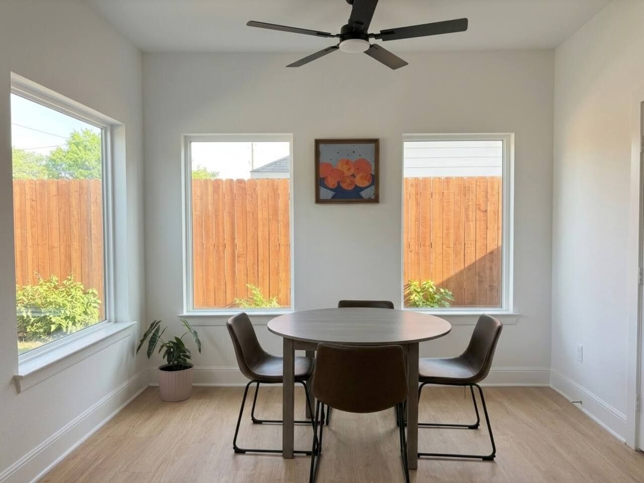 A dining area with a round table and four chairs, large windows with a view of a wooden fence, and a ceiling fan.