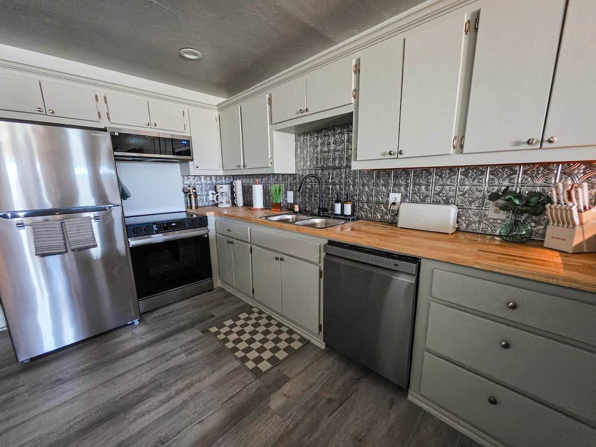 A modern kitchen with stainless steel appliances, light-colored cabinets, a wooden countertop, and a patterned backsplash.