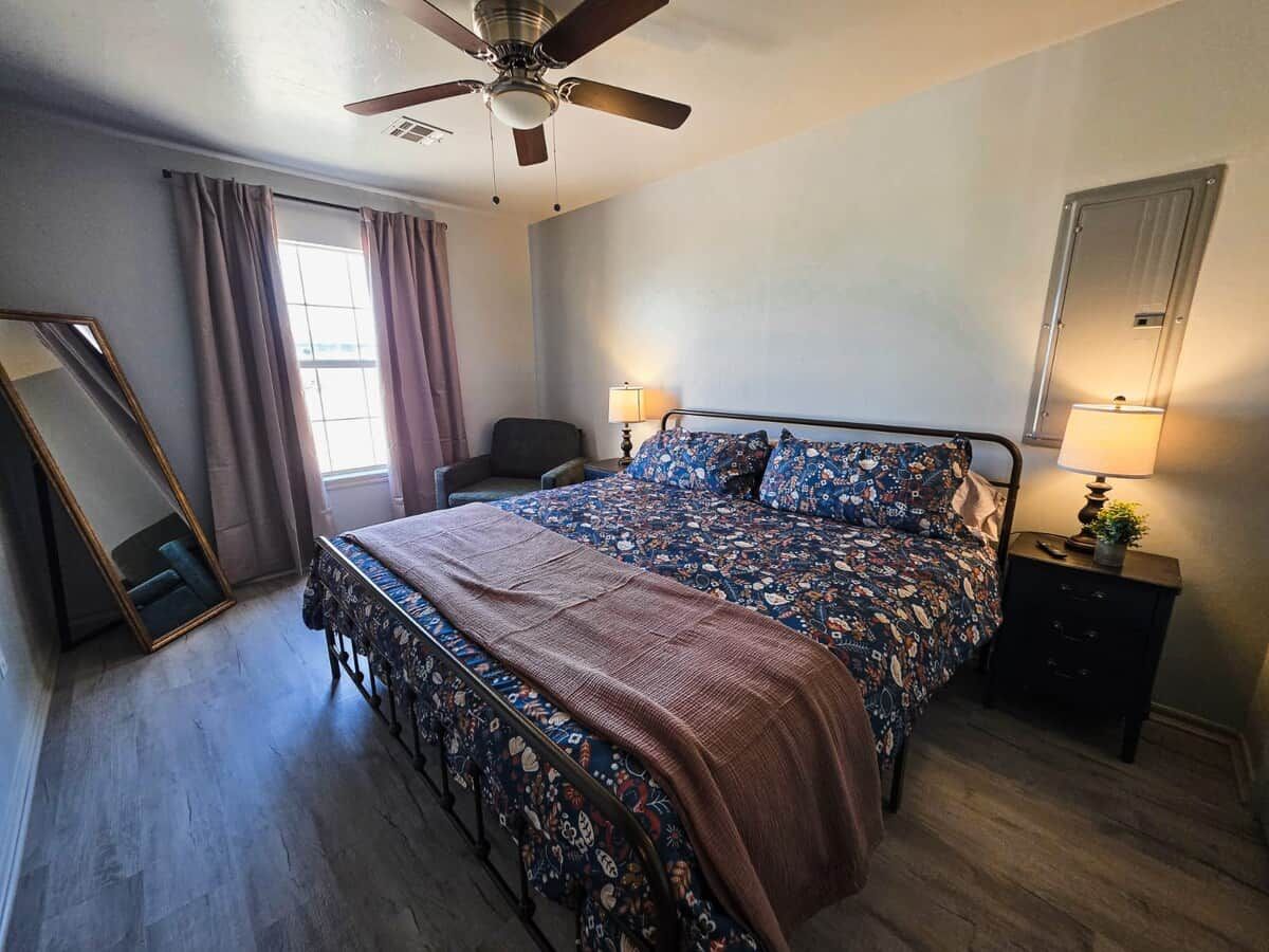 A bedroom with a bed, a nightstand, a mirror, a ceiling fan, and a window with curtains.