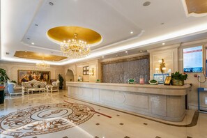 Un vestíbulo de hotel con un mostrador de recepción, candelabros, una alfombra estampada y un mural grande en la pared.