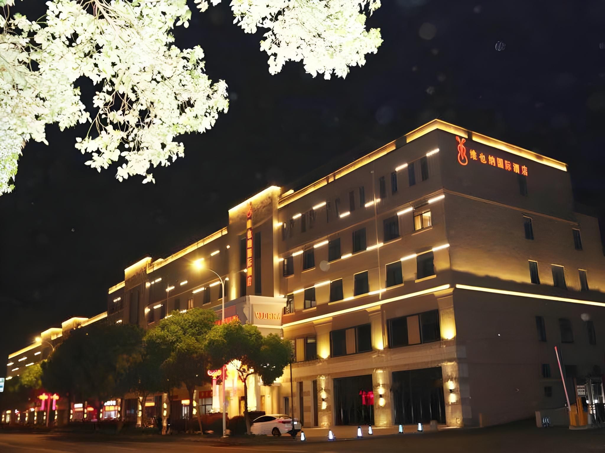 A multi-story hotel building with illuminated signage, a parking area, and a tree with white flowers.