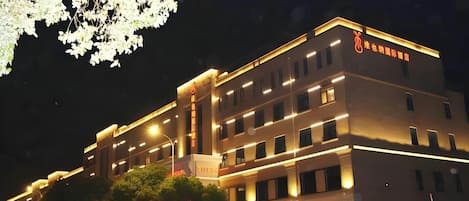 A multi-story hotel building with illuminated signage, a parking area, and a tree with white flowers.