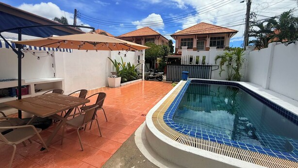 Una piscina con bordo piastrellato, circondata da un patio con sedie e un tavolo, e un'area coperta con luci a stringa.